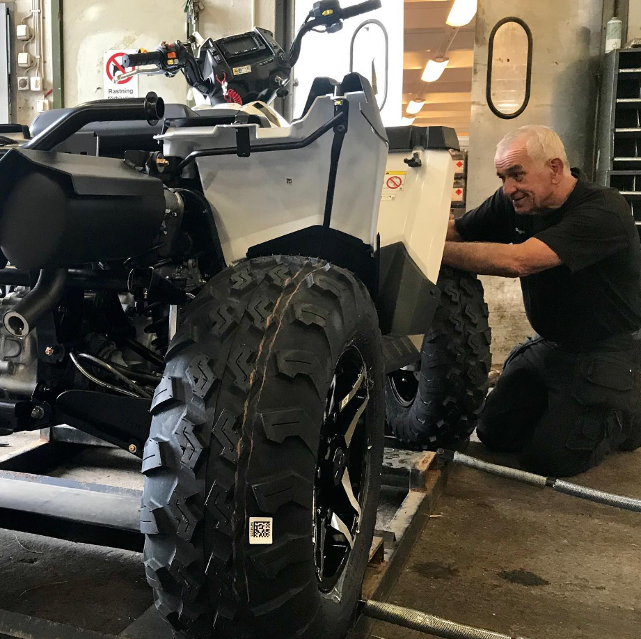 En äldre man arbetar på ett fordon med stora däck, troligtvis en typ av fyrhjuling, i en verkstad eller garage.