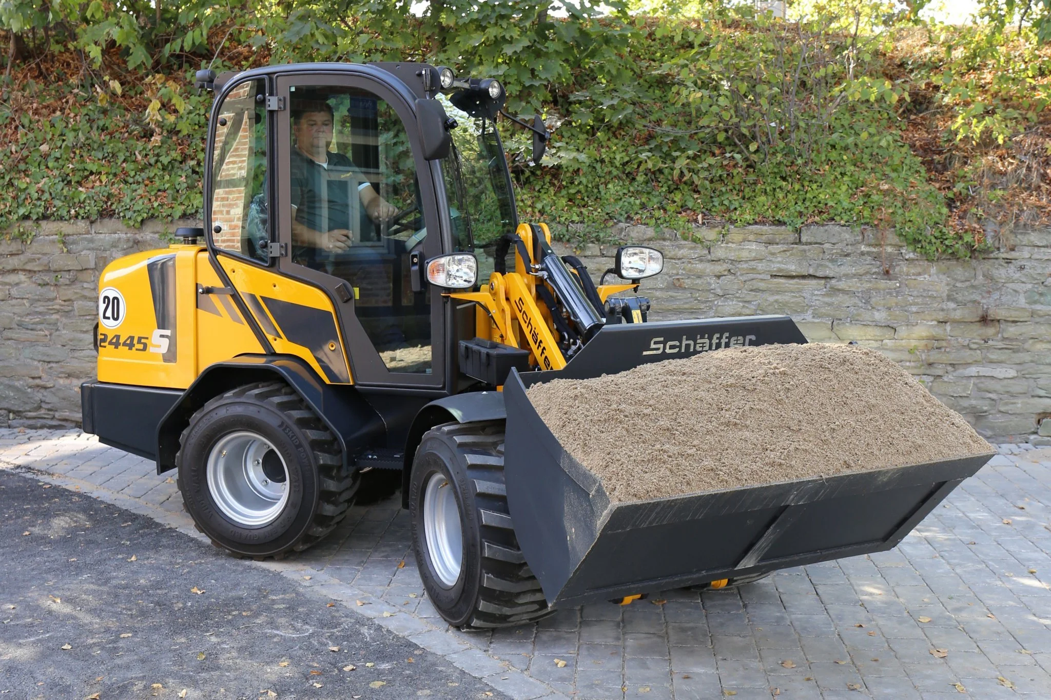 En person kör en liten Schäffer med en frontlastare fyllt med sand, på en stenbelagd yta mot en bakgrund av buskar och en stenmur.