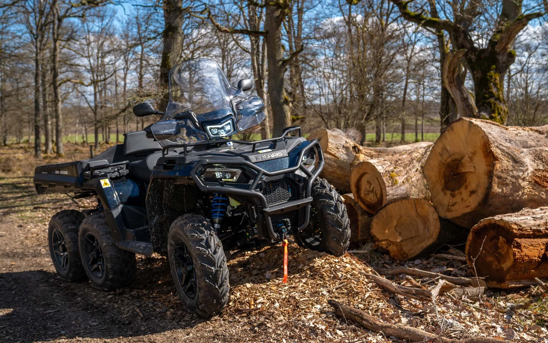 En svart Polaris Sportsman 570 6x6 står bredvid en hög med avhuggna träd på en skogsslinga, under en klar blå himmel.