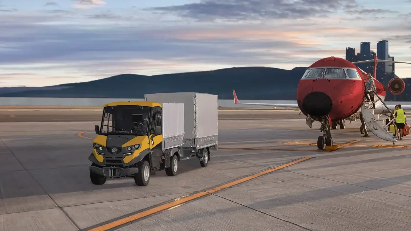 Ett flygplans nosvinge med en röd fenstrale och tillhörande trappa för passagerare med personal i gule västar, samt en elektrisk Alkè på en flygplats bassäng, med berg i bakgrunden, under en molnig himmel.