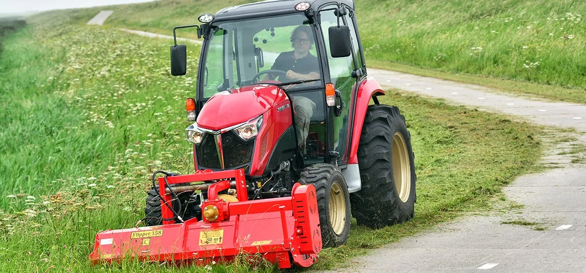 En person kör en röd Yanmar traktor med gräs- och blomgard på ett gräsfält bredvid en grusväg.