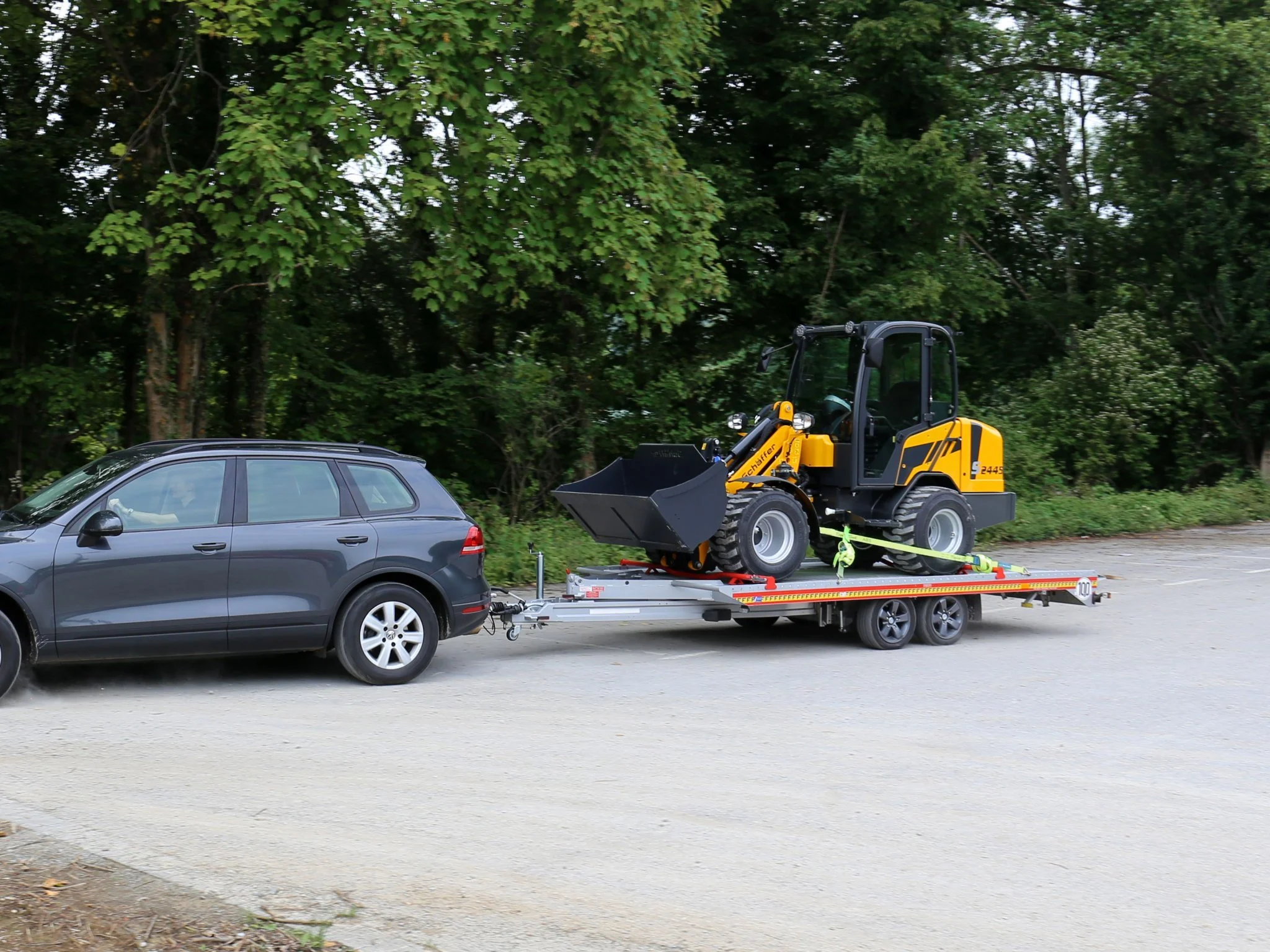 En svart bil är kopplad till en trailer som transporterar en liten gul Schäffer på en parkeringsplats med grönskande träd i bakgrunden.