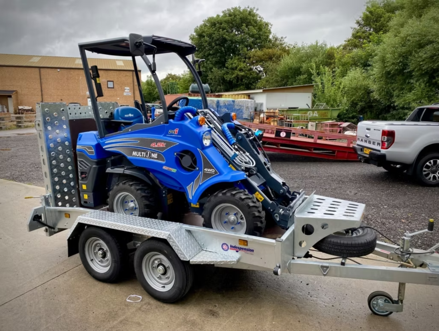 En liten blå MultiOne på en transportvagn utomhus, med ett lager av träd och en byggnad i bakgrunden.
