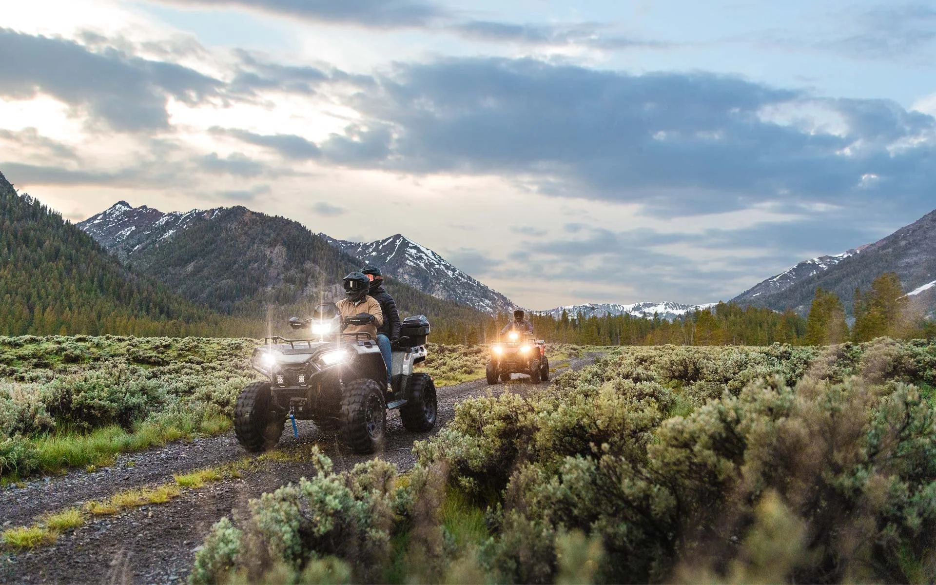 Två personer åker på Polaris Sportsman 570 längs en grusväg i en bergig, grön och snöbeklädd landskap, under en molnig himmel.