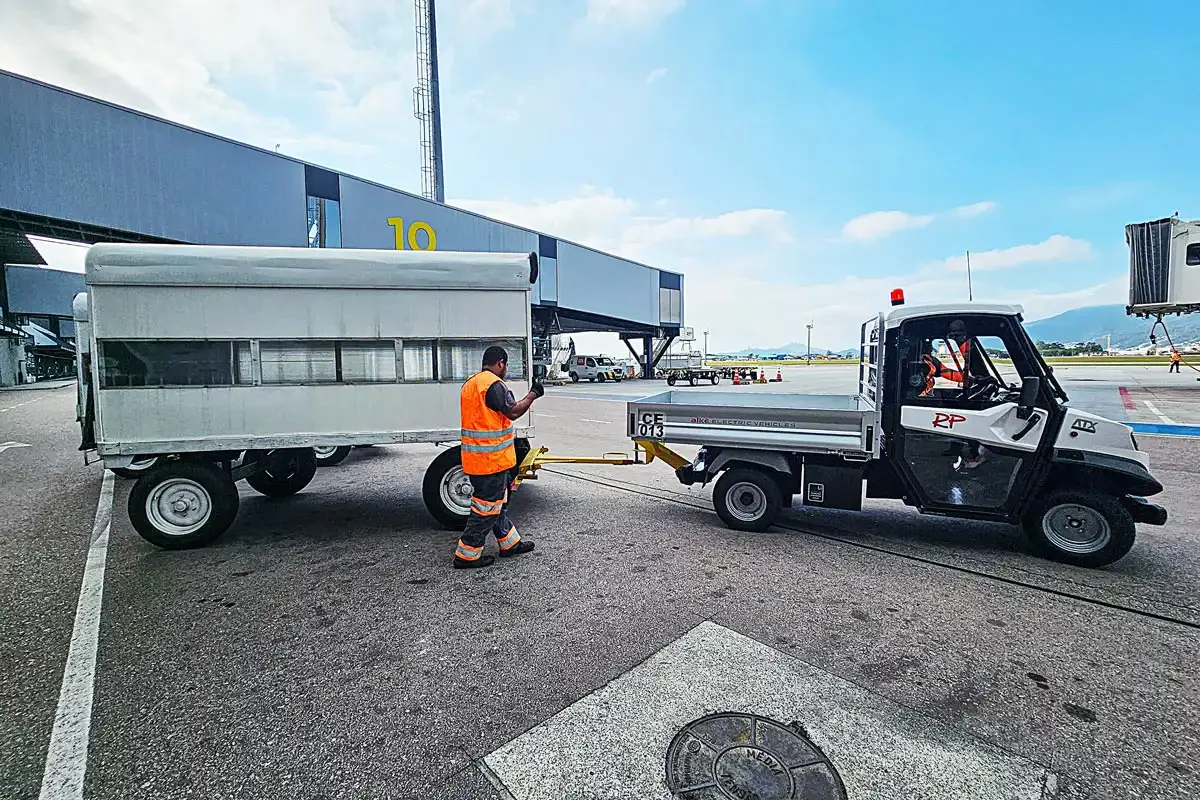 En person i orange säkerhetsväst kopplar en transportvagn till en liten elektrisk Alkè vid en flygplats.