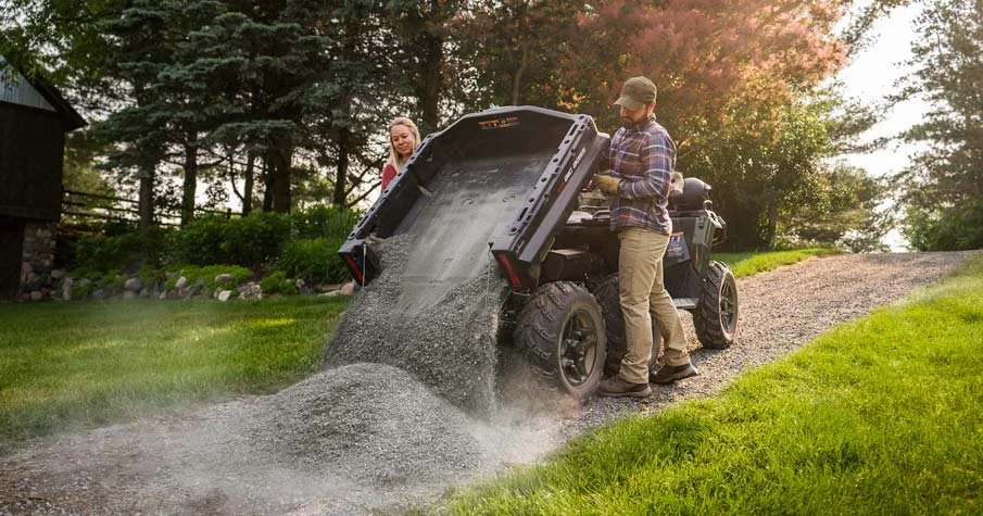 Två personer står utomhus bredvid en Polaris Sportsman 570 6x6  som tömmer ut en mängd grus på grusvägen.