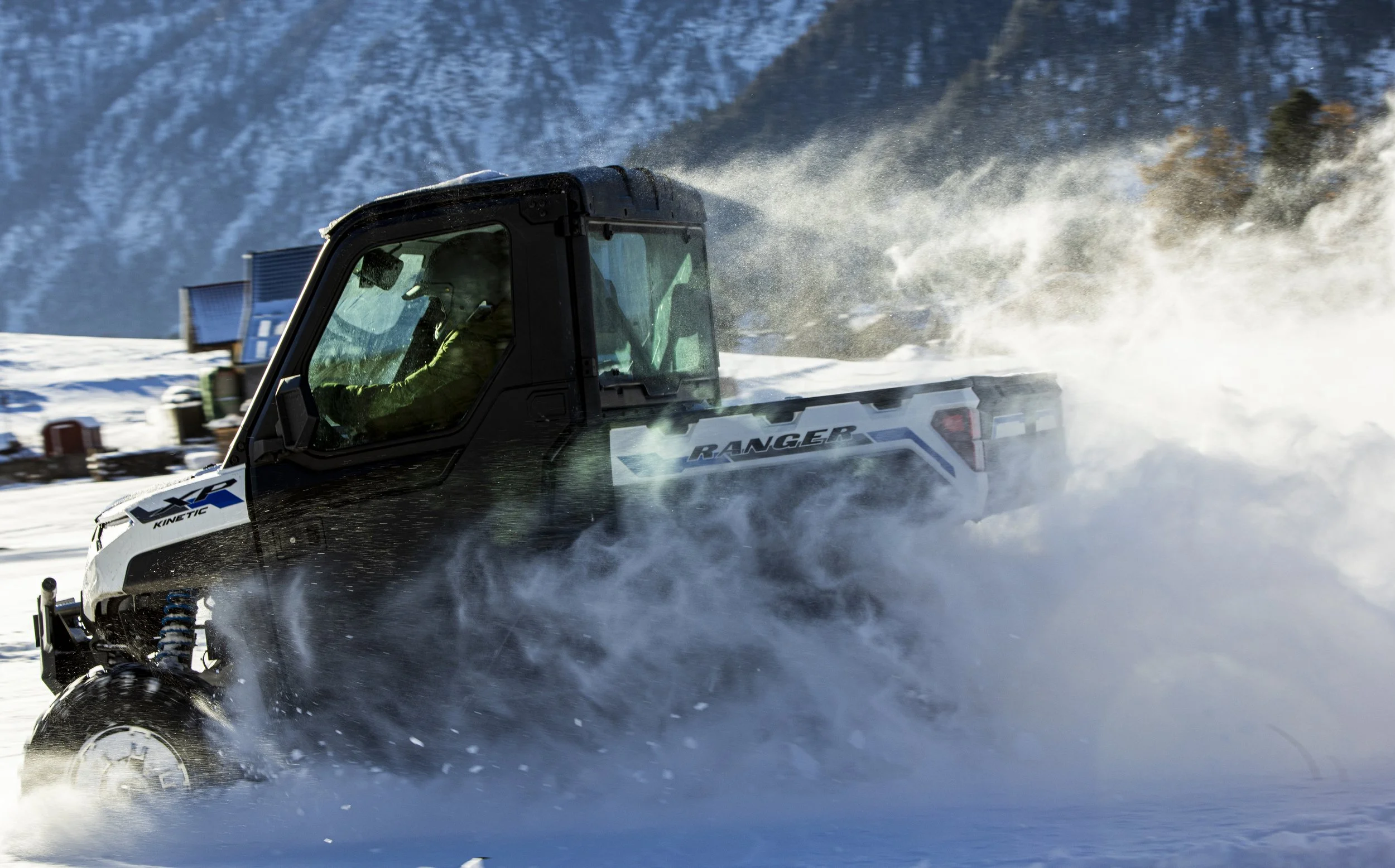 En fyrhjuling som kör genom snö, med snön som sprutar upp runt den, i ett vintrigt landskap med snötäckta berg i bakgrunden.