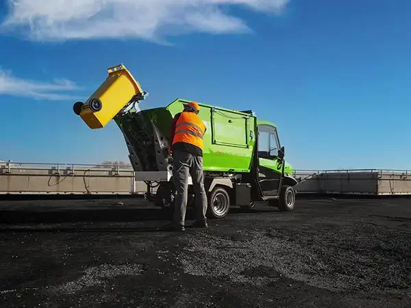 En person i orange väst som tömmer ett gulaktigt soptuna i en grön Alkè  på en väg under en klar blå himmel.
