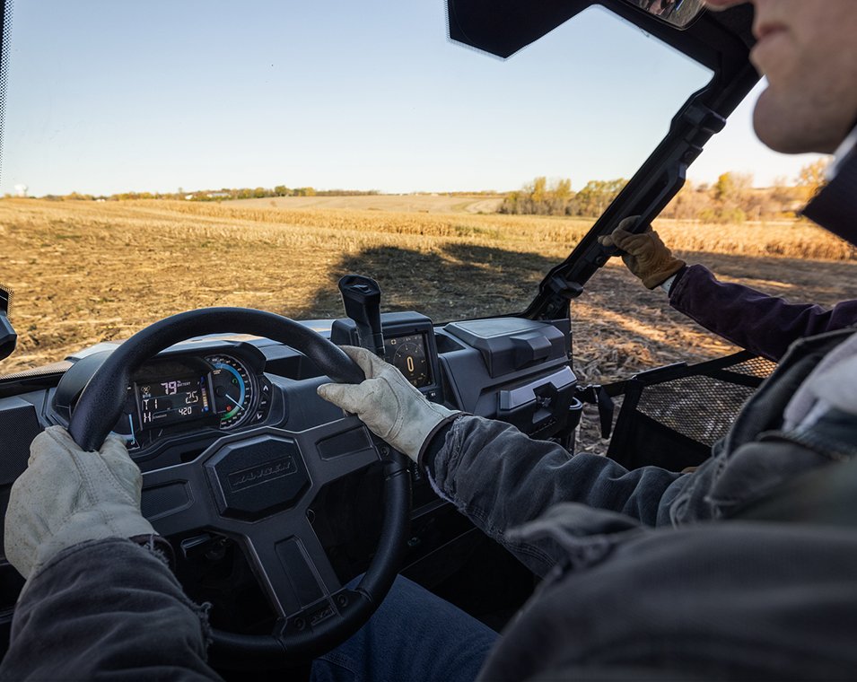 Två personer som kör en Polaris Ranger XP Kinetic på ett fält eller jordbruksmark, med panoramavy av landskapet i bakgrunden.