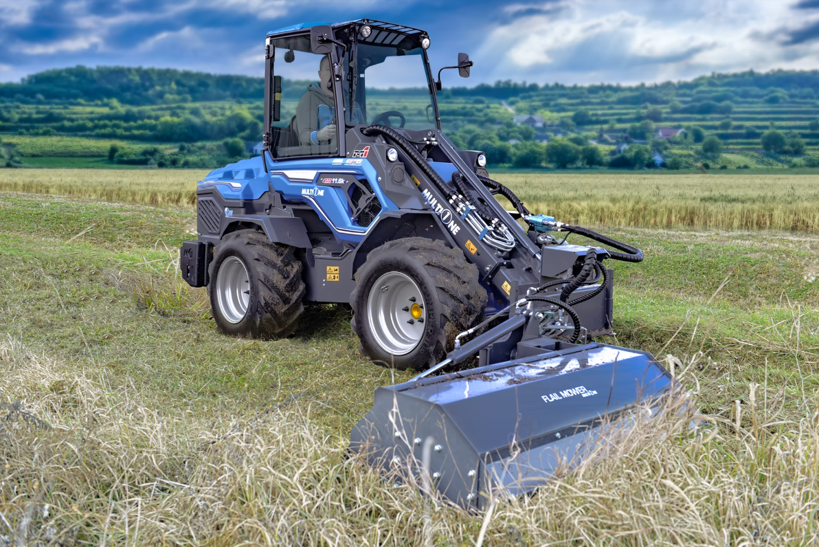 En MultiOne med gripredskap som arbetar på ett fält med gräs eller skörd. Landskap med gröna kullar och ett molnigt himmel i bakgrunden.