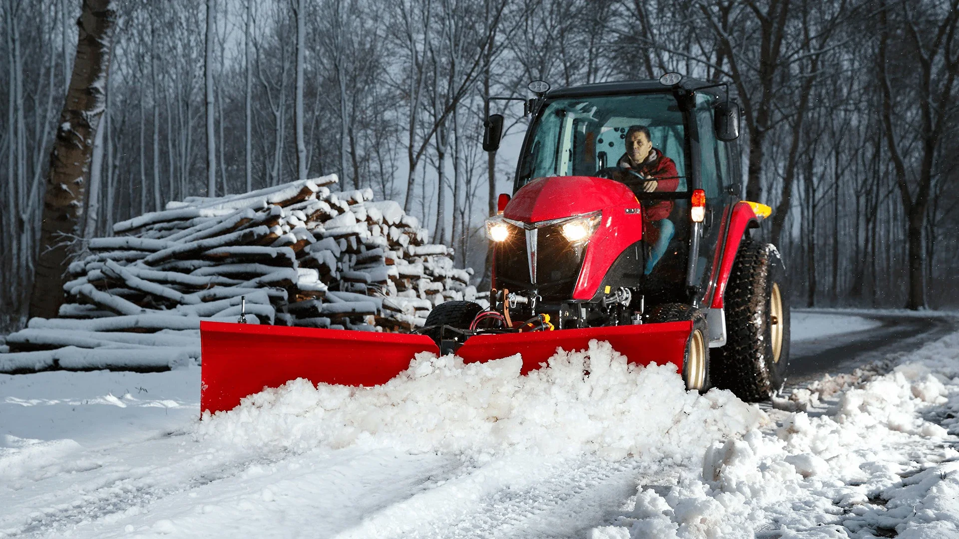 En person kör en röd Yanmar på en snöig skogsväg under dagtid. Det finns ett högt högt lager av snö och en hög stapel av ved på vänster sida av vägen.