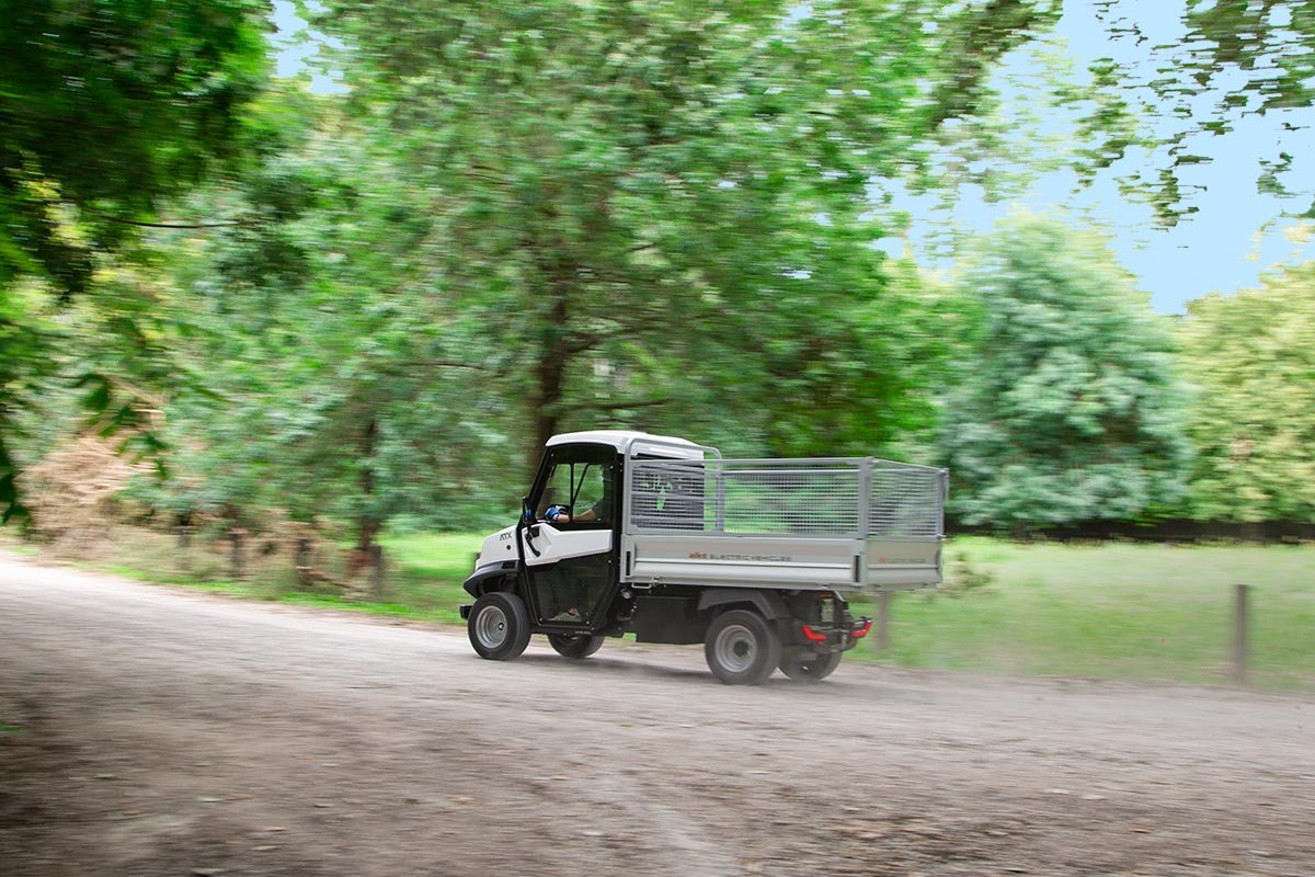 En liten elektrisk Alkè kör på en grusväg genom en skog med gröna träd och löv.