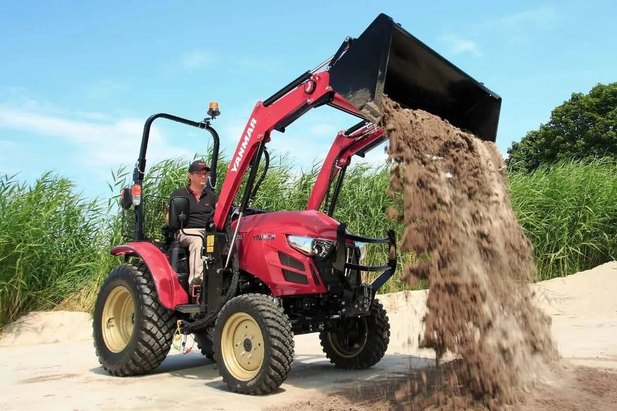 En person sitter på en Yanmar som tömmer jord ur en skopa framtill på ett sandigt område med gröna gräs och träd i bakgrunden.