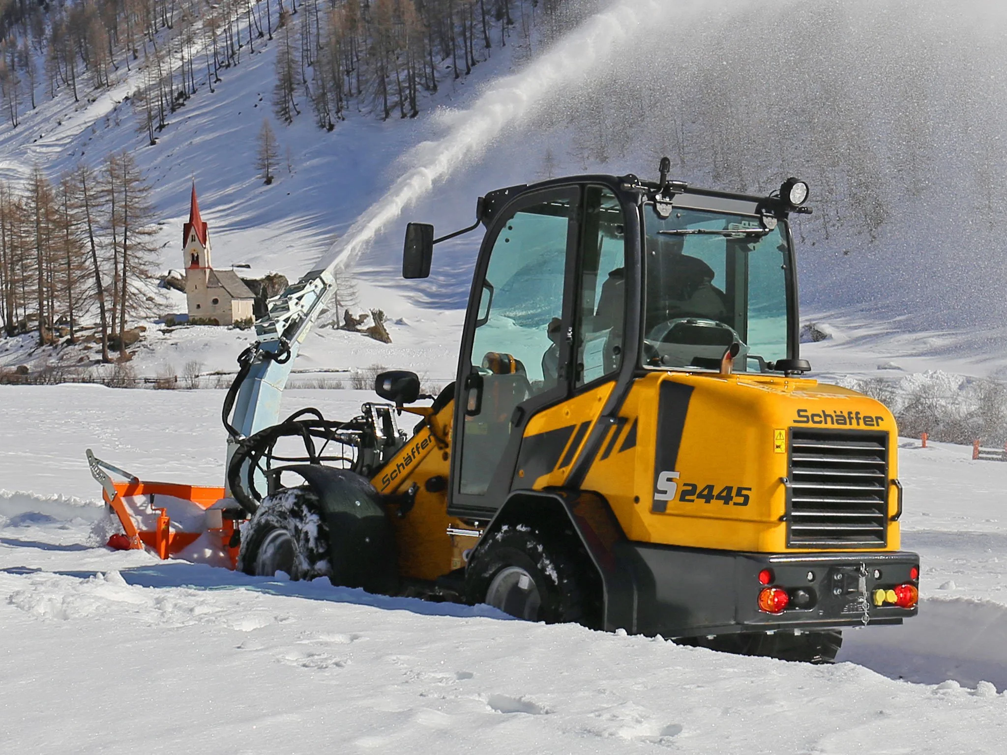 En gul Schäffer i snötäckta landskapet, bakgrund med snöklädda kullar och en liten kyrka.