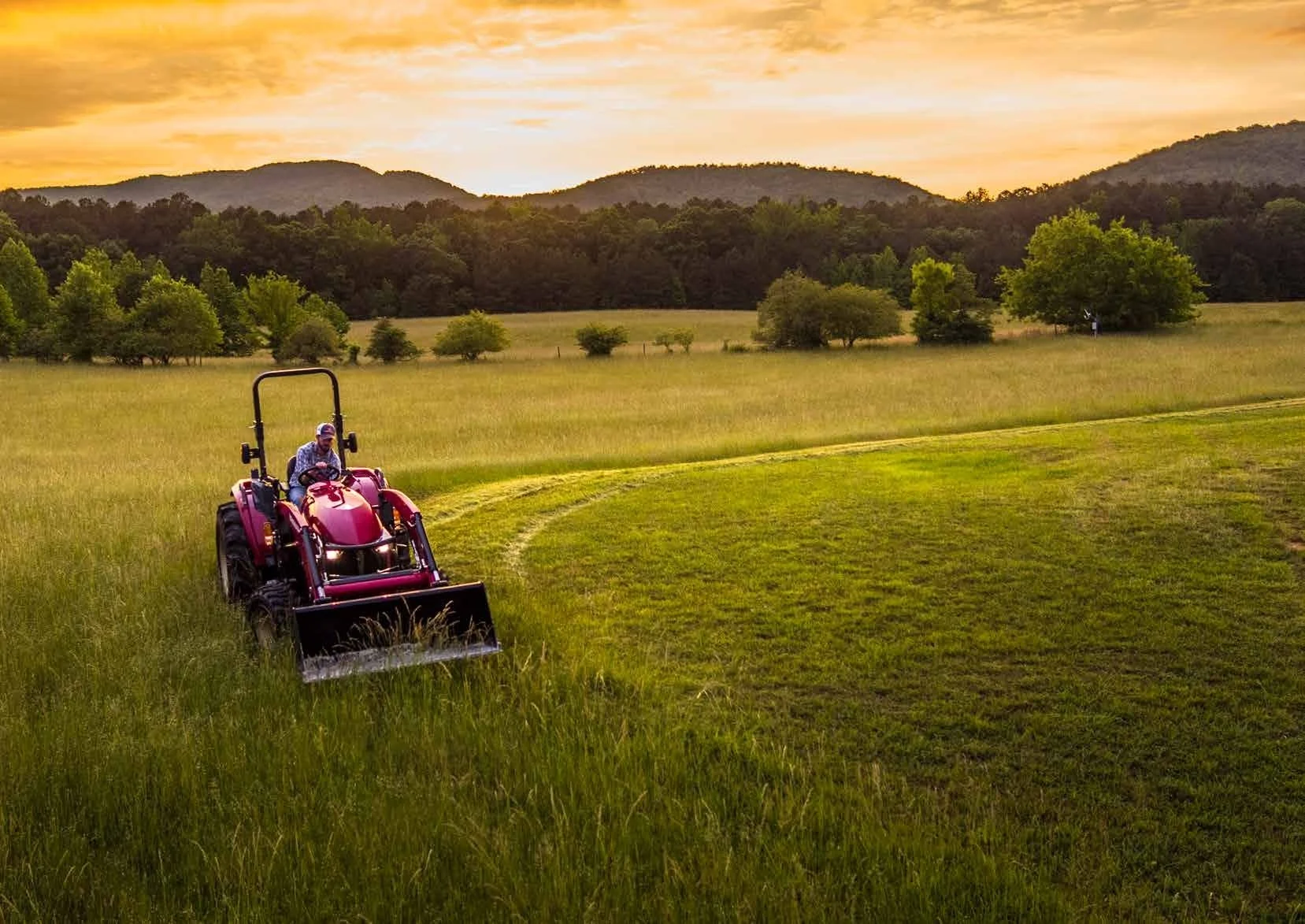Man som kör en röd Yanmar traktor på en gräsmatta i ett landskap vid solnedgången, med träd och kullar i bakgrunden.