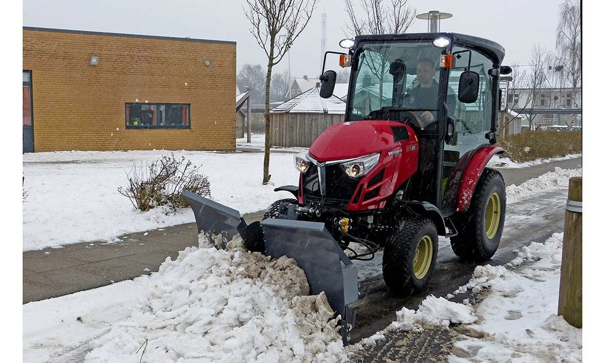 En person kör en Yanmar snöslunga på en trottoar under vinterväder, omgiven av snö och några träd.