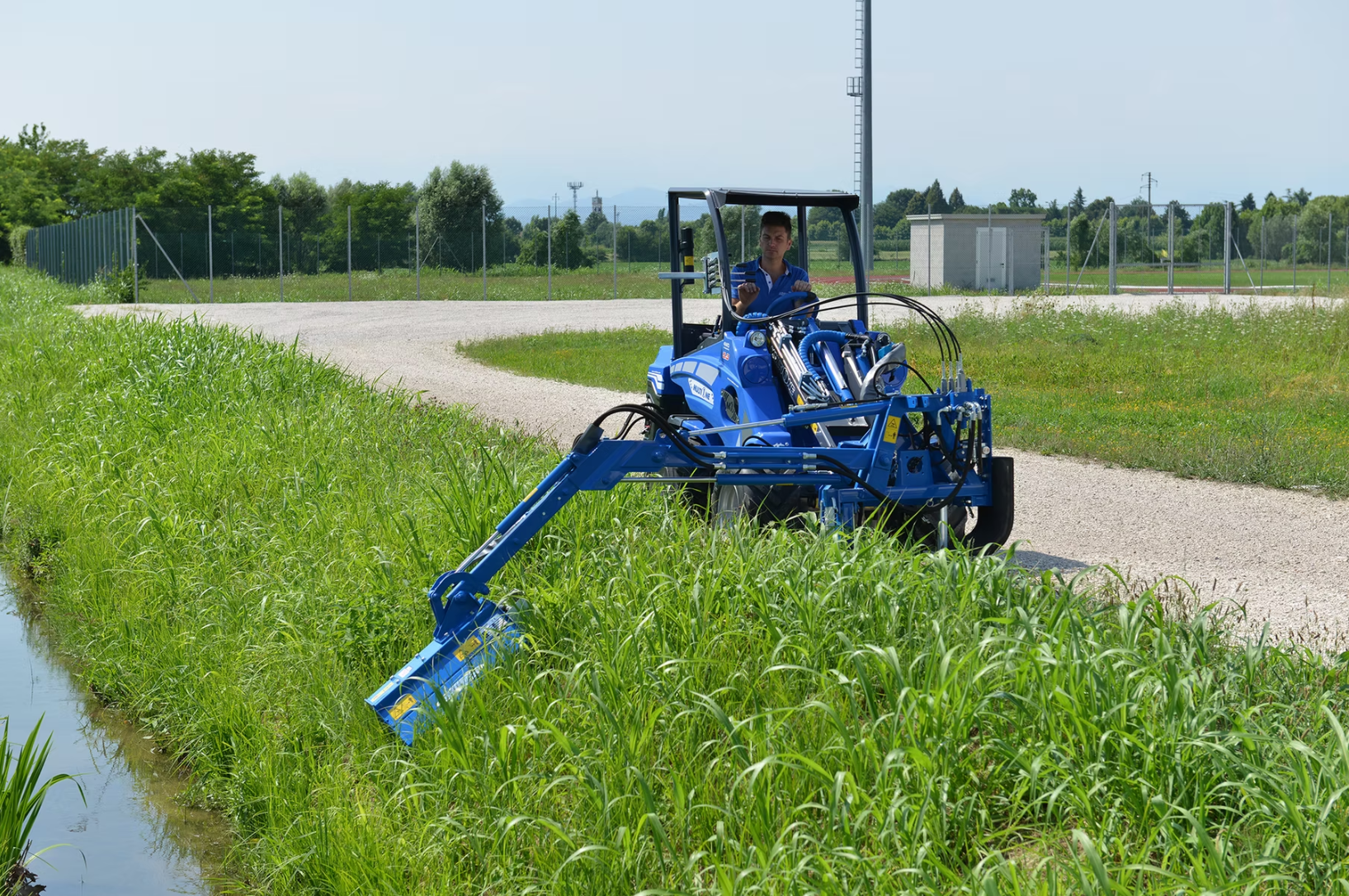 En man som kör en MultiOne på en gräsmatta bredvid en väg i ett sommarlandskap.