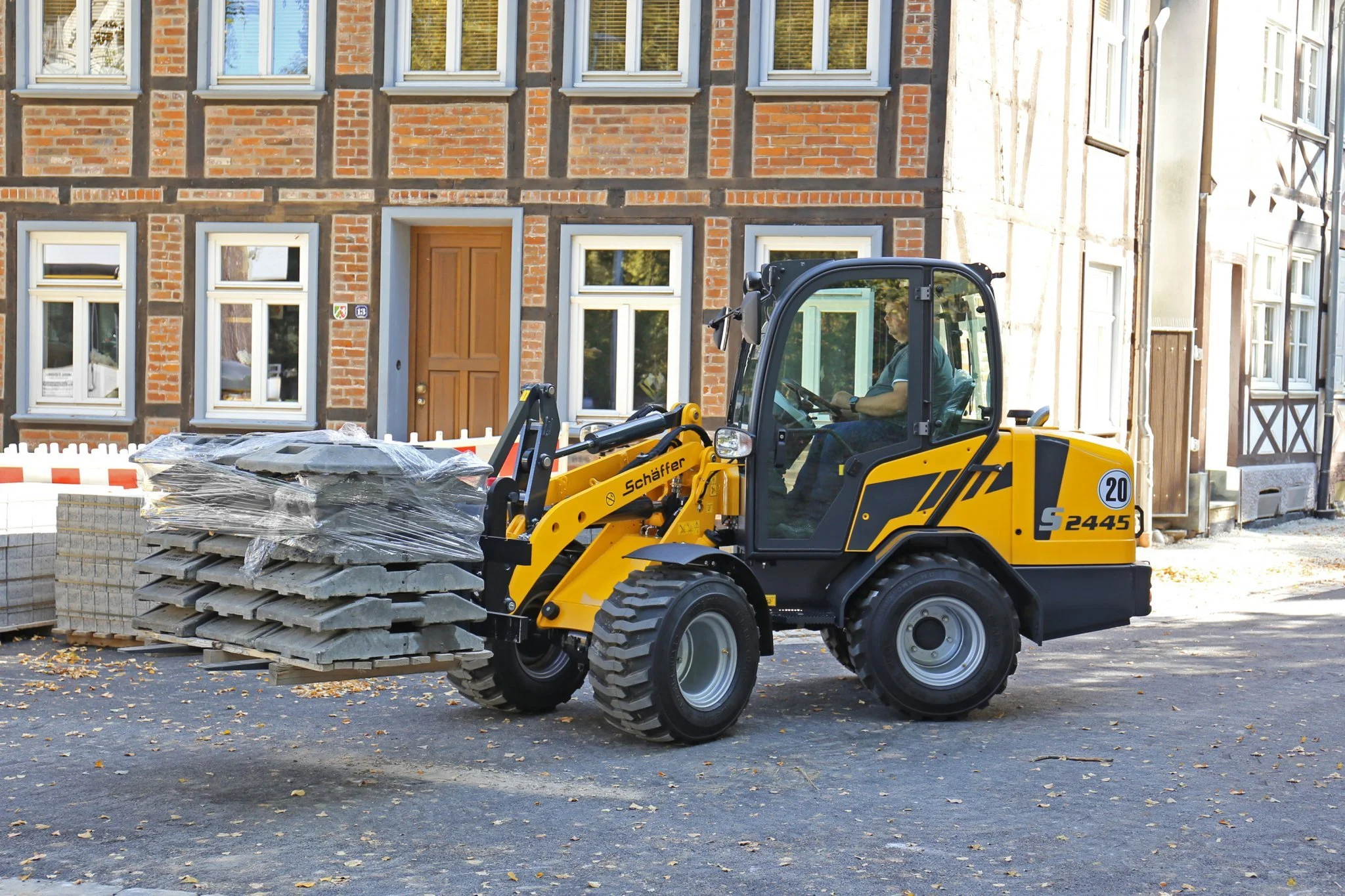 En gul Schäffer lastar stenblock i stadsmiljö med äldre tegelhus i bakgrunden.