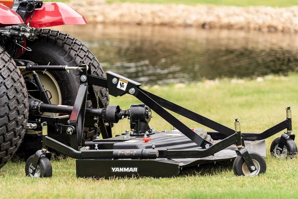 En gräsklippare med stora bakhjul och mindre framhjul, kopplad till en Yanmar traktor, på gräsnära område vid en kropp av vatten.