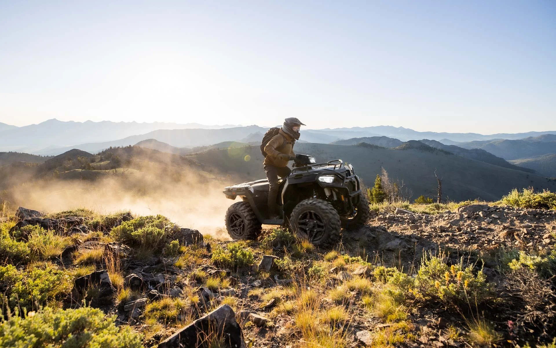 Person riding a Polaris Sportsman 570 on a rocky trail in a mountainous landscape with green vegetation.