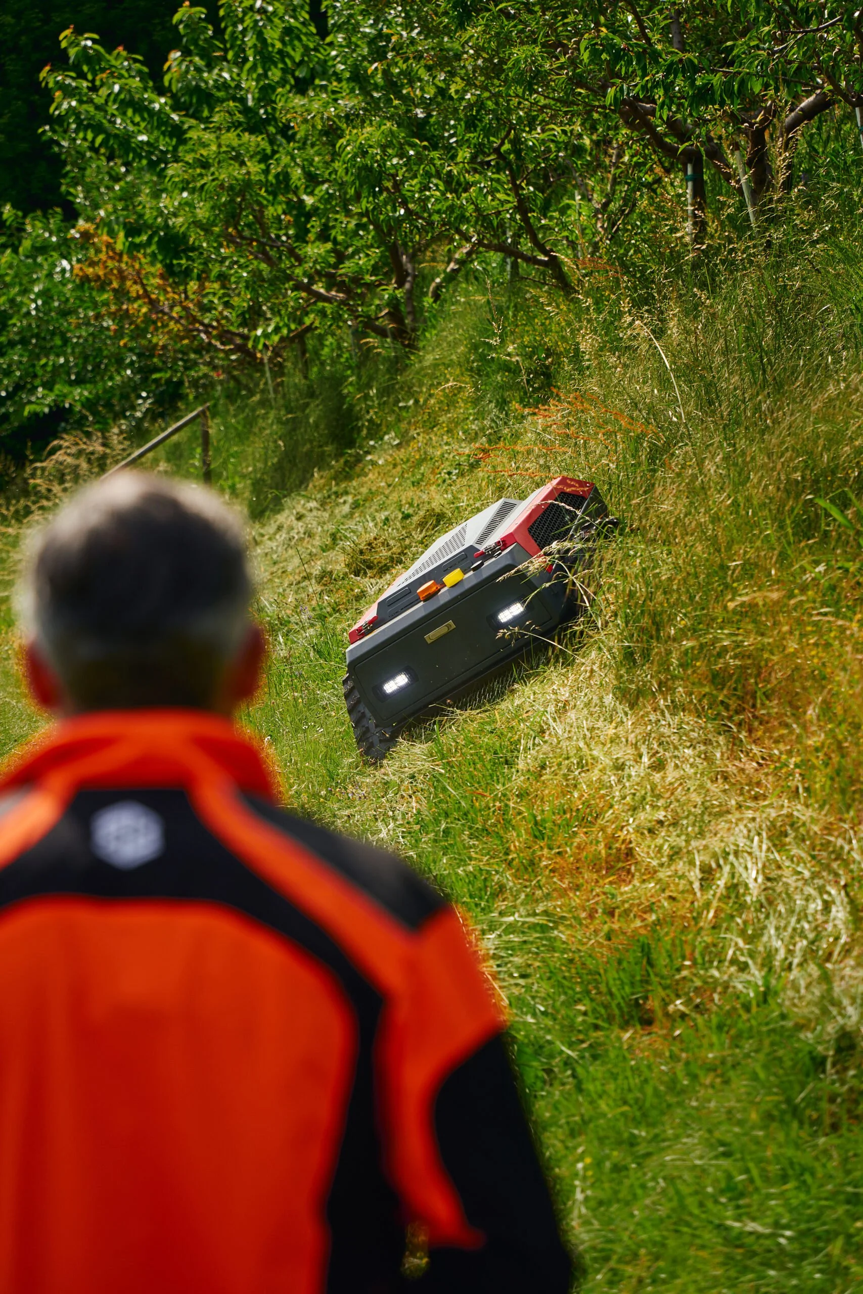 En person i en orange och svart jacka tittar på en BlueBird Fjärrstyrd Släntklippare i ett grönt fält med träd i bakgrunden.