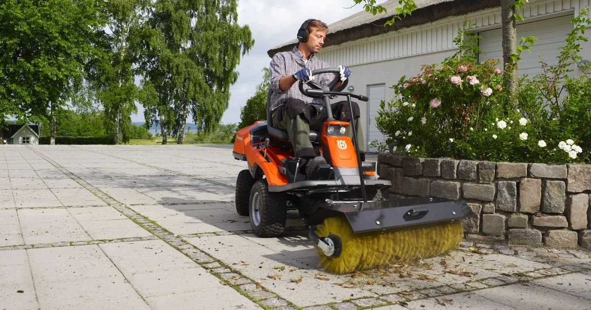 En man som kör en Husqvarna Rider på en uteplats med stenläggning, nära en blomsterlåda med rosa och vita blommor.