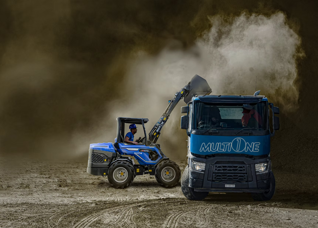 En person i en blå MultiOne som använder en skopor för att flytta jord till en lastbil märkt 'MULTIONE' på en byggplats.