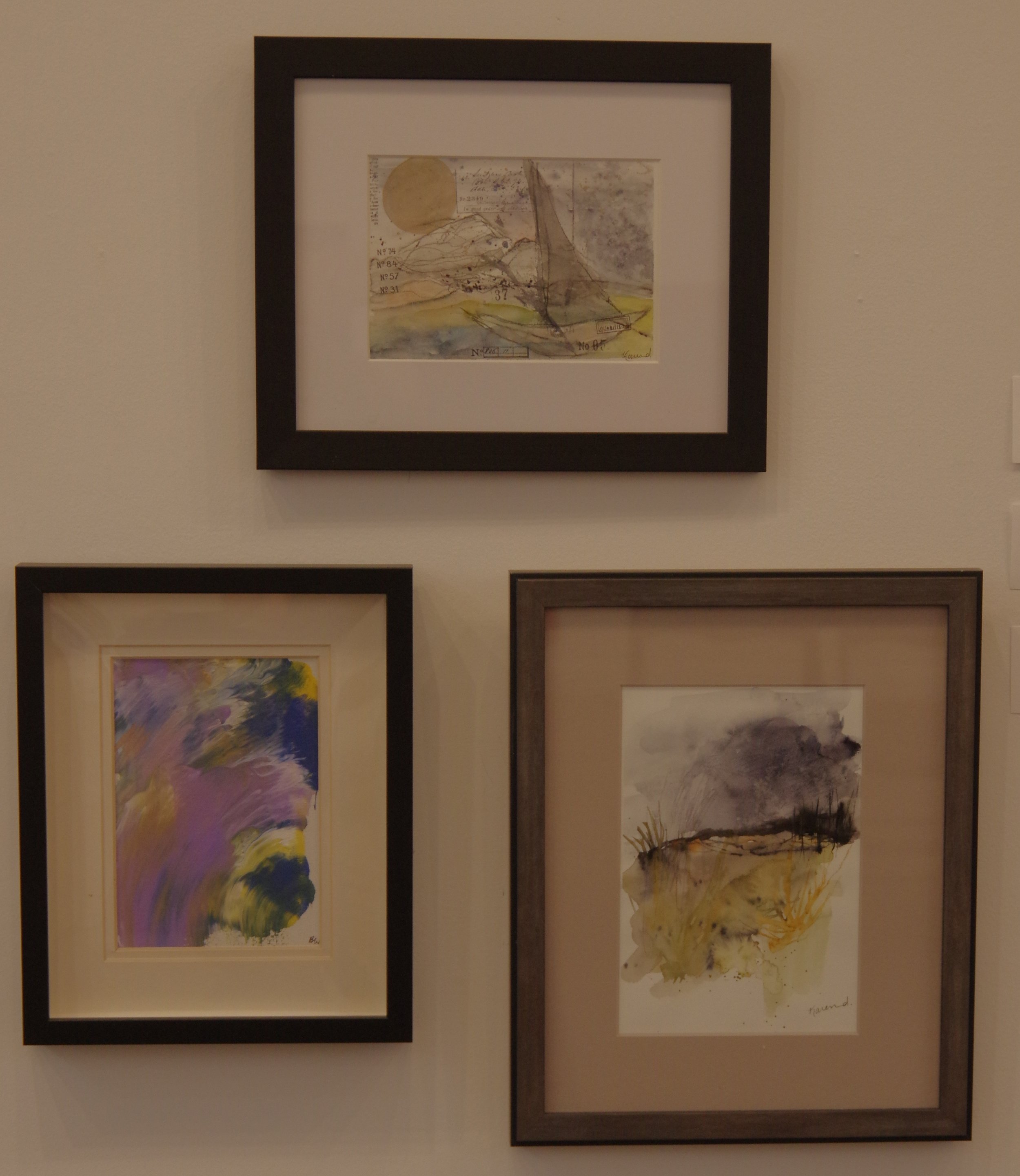 Three framed watercolor paintings hanging on a beige wall, with the top frame containing a sketch of a sailboat on water with a golden sun in the background, the bottom left frame showing an abstract swirl of purple, yellow, green, and blue, and the bottom right frame depicting a landscape with cloudy sky and trees in muted colors.