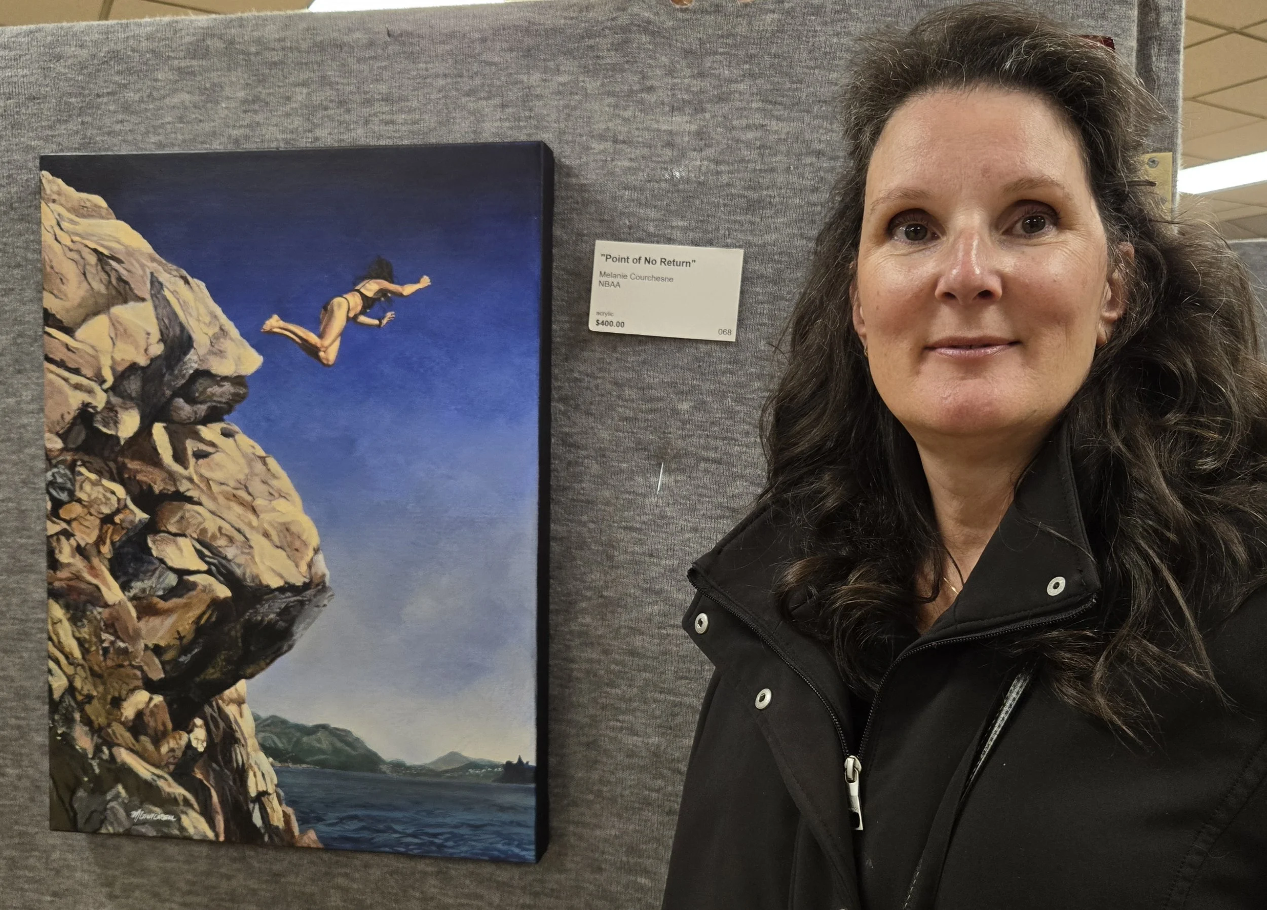 A woman with dark curly hair and wearing a black jacket standing next to a painting of a cliff and a person diving into water, displayed at an art gallery.