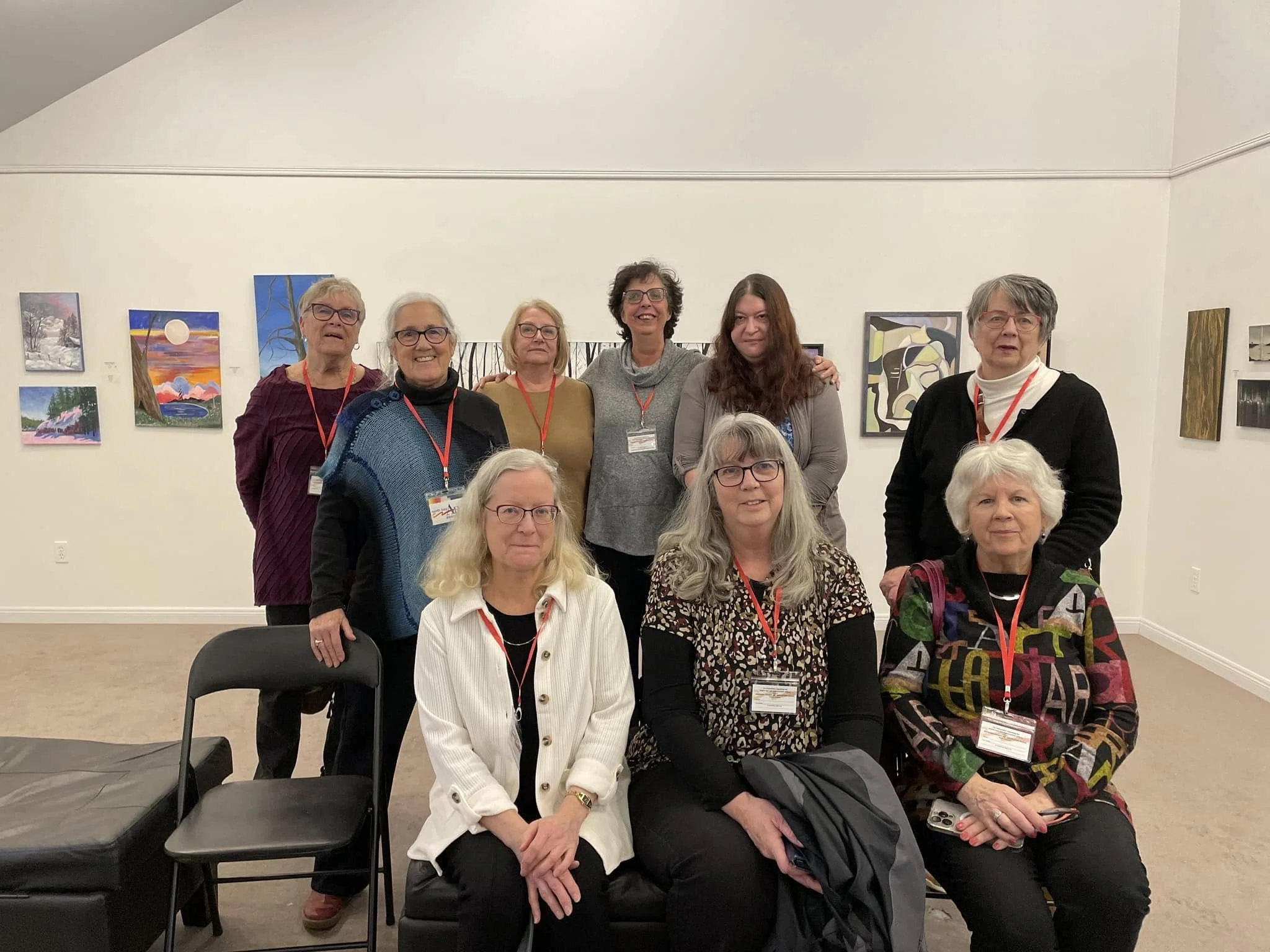 Group of nine women in an art gallery posing for a photo, with colorful paintings on the wall behind them.