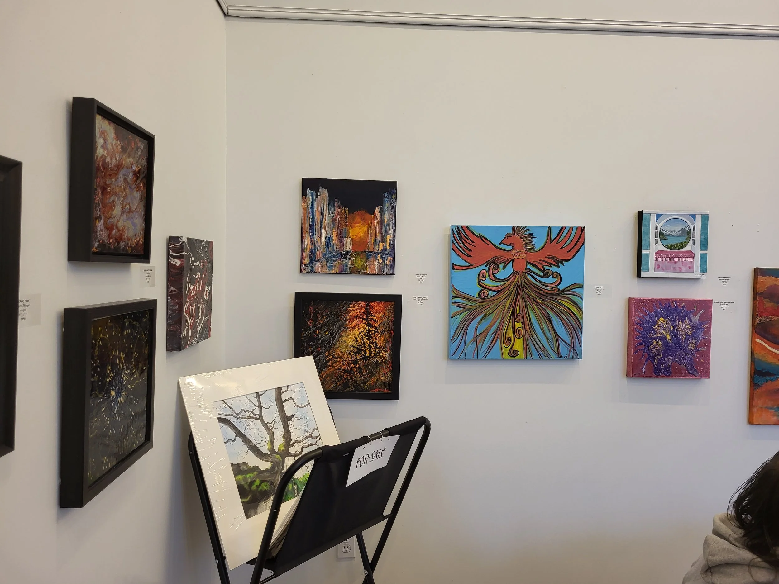 Gallery wall with multiple colorful abstract paintings, one of which features a prominent bird figure, and a framed piece with a tree, with a sign indicating for sale.