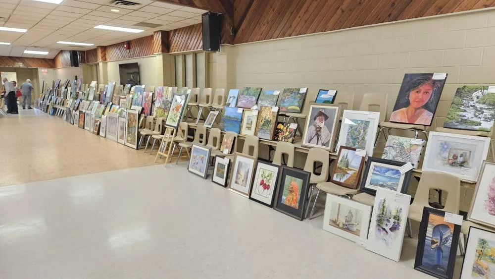 Art display at an indoor gallery or event, with paintings leaning against chairs and the wall, featuring landscapes, portraits, and abstract art.