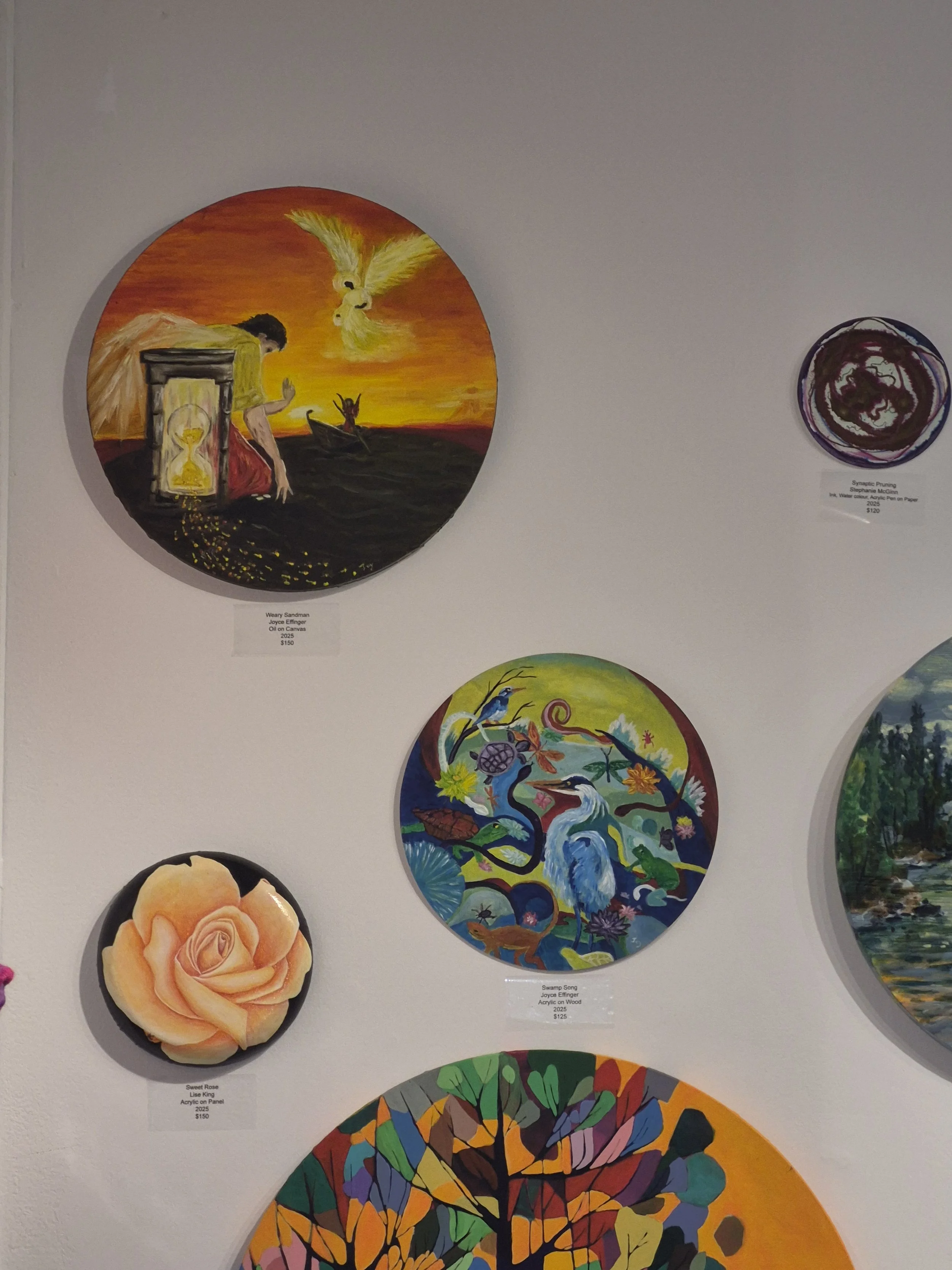 Art exhibit featuring colorful paintings, including a circular painting of a woman with a bird, a dragon in a pond, a pink rose, and abstract designs.