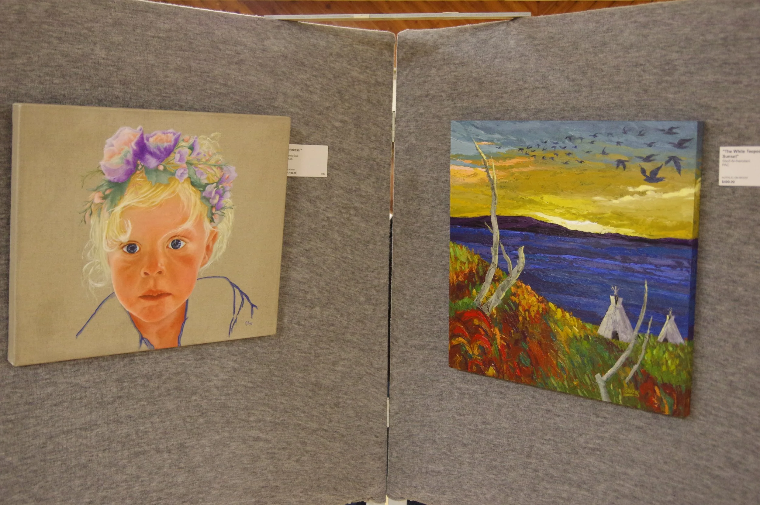 Two paintings displayed on a gray fabric-covered partition. The left painting is a realistic portrait of a young girl with blond hair, blue eyes, and a flower crown of purple and pink flowers. The right painting is an impressionistic landscape showing a vibrant sunset over water with trees and small white teepees in the foreground.