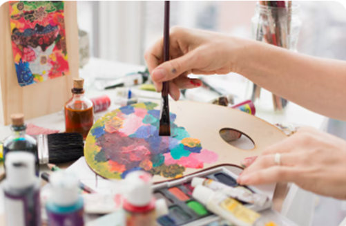 Close-up of a person's hands painting on a colorful palette in an artist's studio with paints and brushes around.