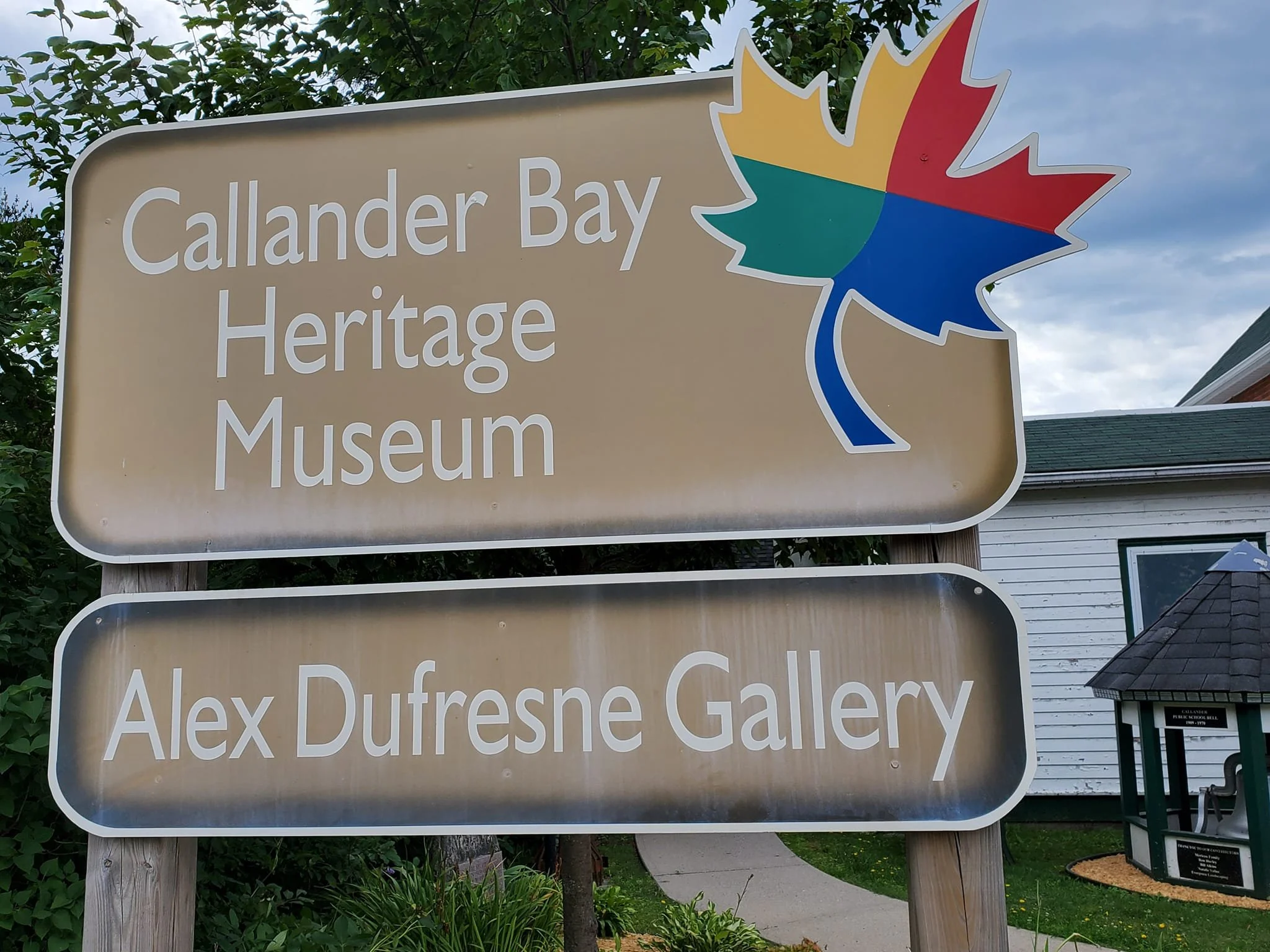Sign for Callander Bay Heritage Museum and Alex Dufresne Gallery with a colorful maple leaf logo, outdoors with a house and trees in the background.