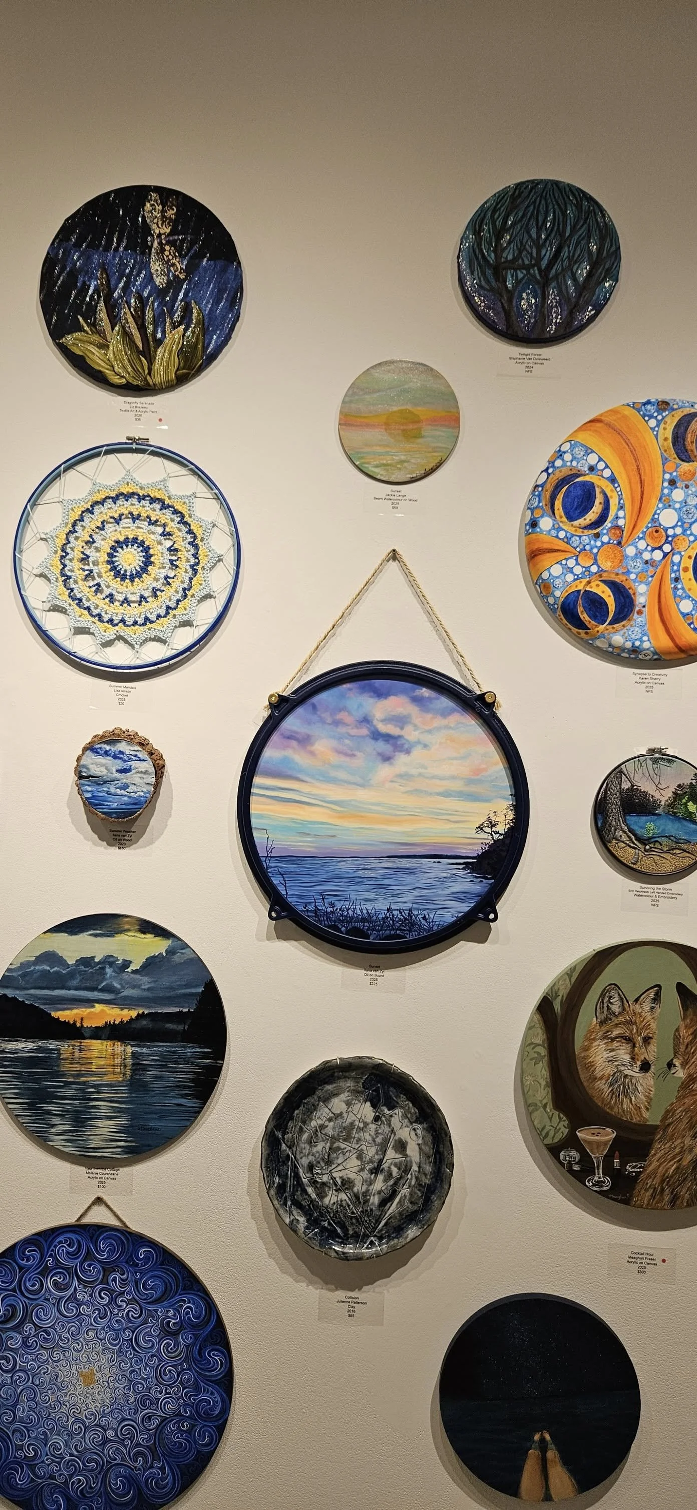 A wall display of various round painted artworks, including landscapes, abstract designs, and nature scenes, arranged in a gallery setting.