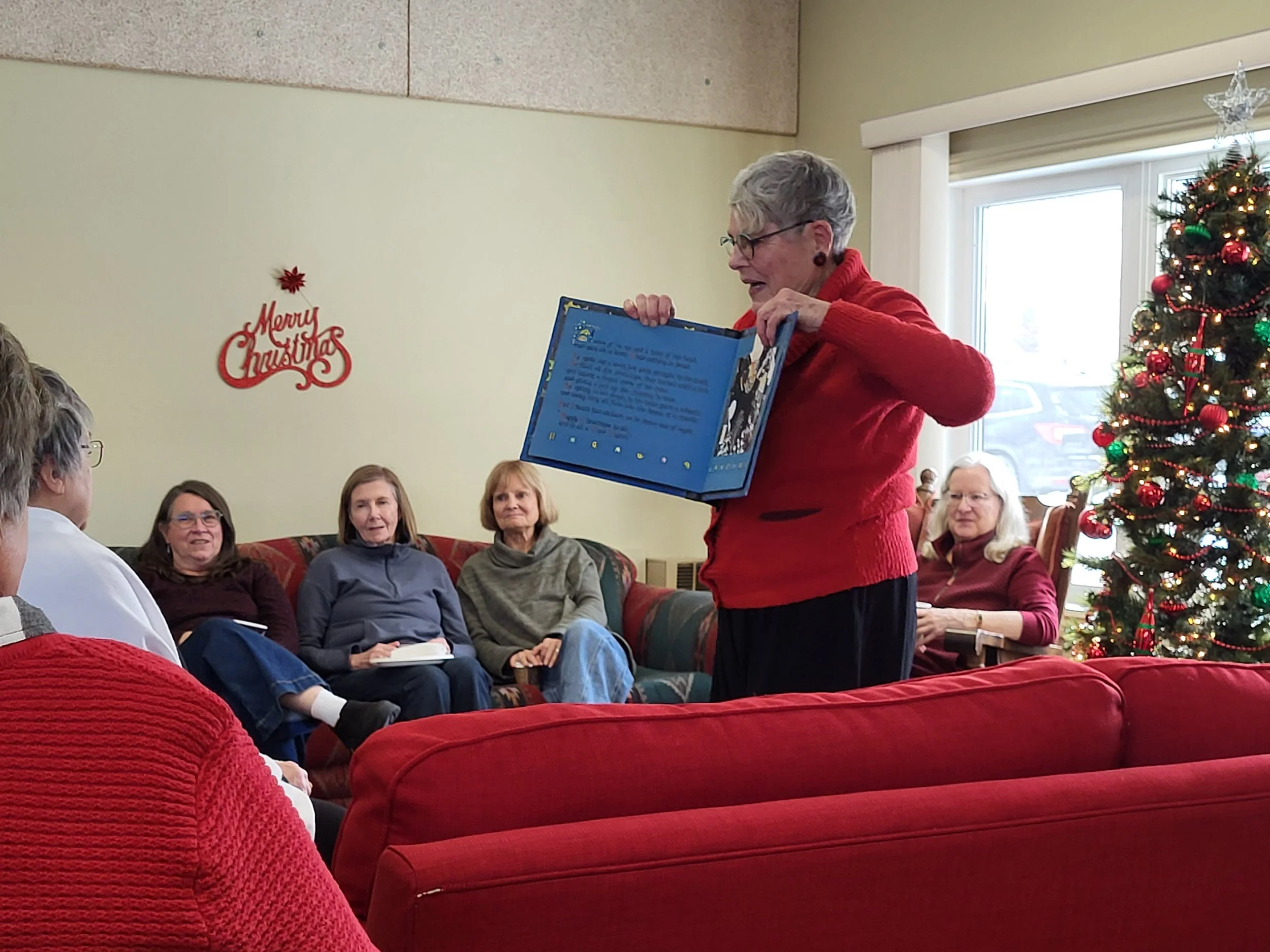 A group of women gathered in a living room decorated for Christmas, with a Christmas tree. An elderly woman in a red sweater is showing a large blue book to the group.
