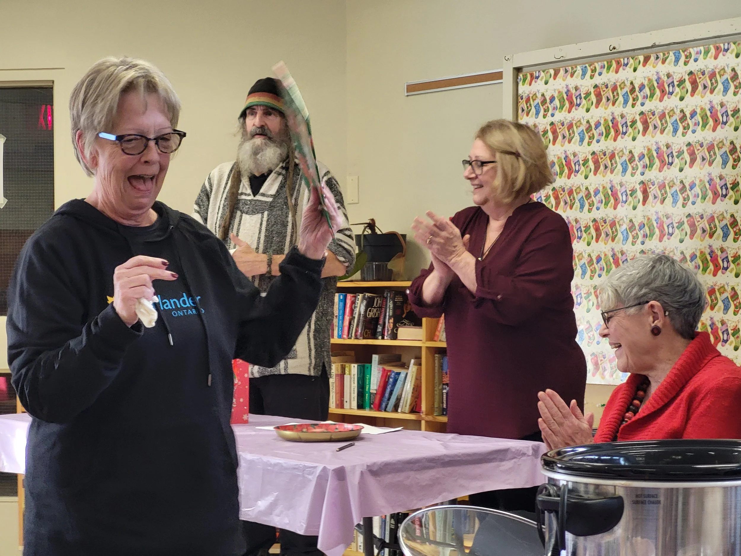 A group of four women celebrating, with one woman holding a wrapped gift, in a room decorated with colorful Christmas stockings on the wall.