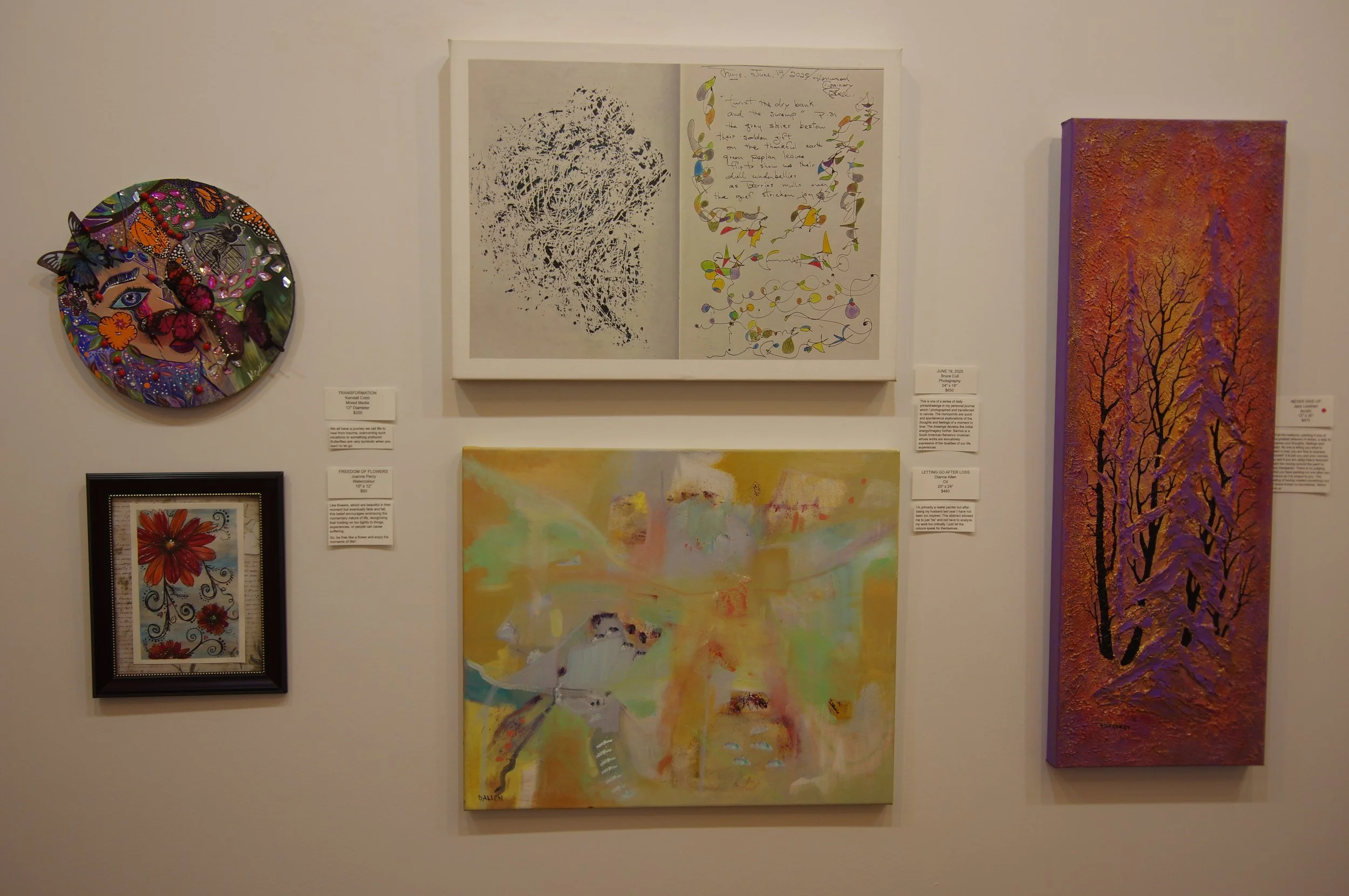 Art display with five pieces: a colorful circular mixed media piece with butterflies and an eye, a framed floral illustration with red flowers, a large abstract painting with pastel colors, a white-framed piece with black ink tree and handwritten notes, and a tall, textured vertical painting of a winter scene with trees and a sunset sky.