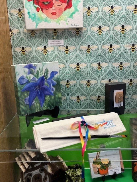 Display of artwork and artistic accessories in a room with bee-themed wallpaper. Features paintings of a woman's face and purple flowers, a black electrical outlet, a small black box, and a table with art tools and a cloth with a rainbow ribbon.