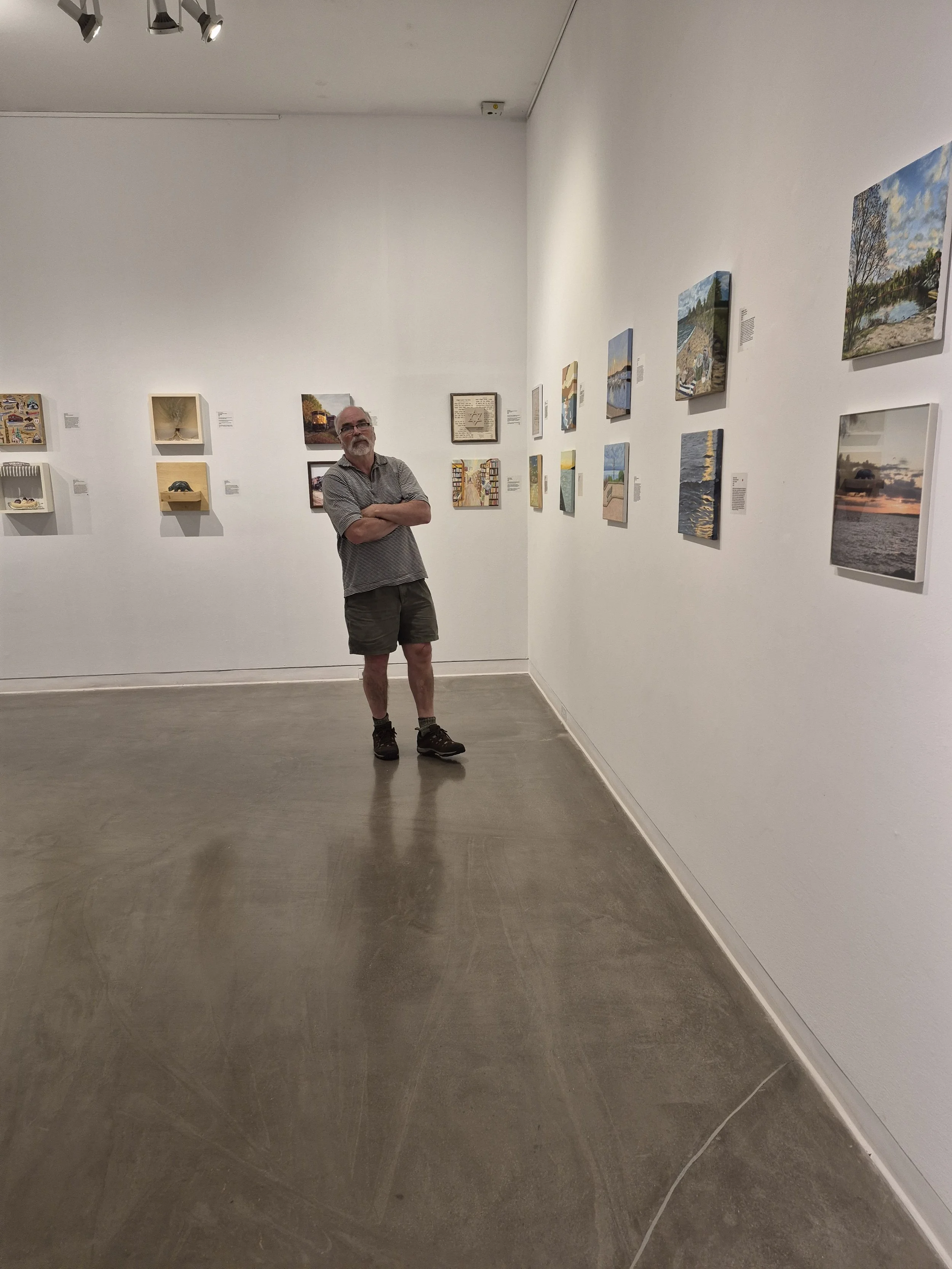 A man with gray hair, beard, and glasses, wearing a patterned short-sleeve shirt, shorts, and hiking shoes, stands with arms crossed in an art gallery, viewing framed paintings and textured artworks on white walls.