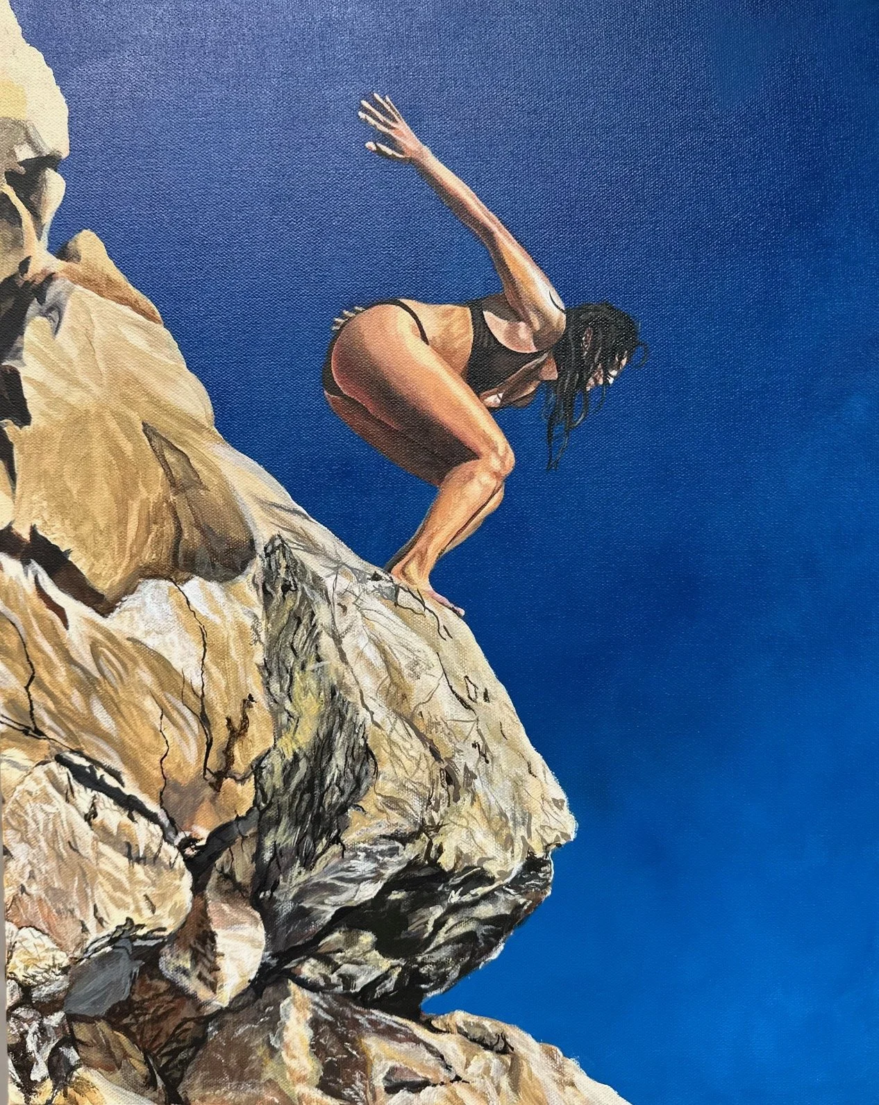 A person with long, dark hair in black swimwear crouching on top of a rocky cliff against a clear blue sky.