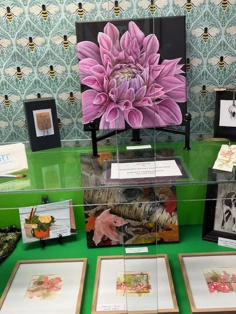 A framed pink flower artwork displayed on a black easel, with several smaller framed nature-themed prints and crafts on a green table, and wallpaper with bee patterns in the background.
