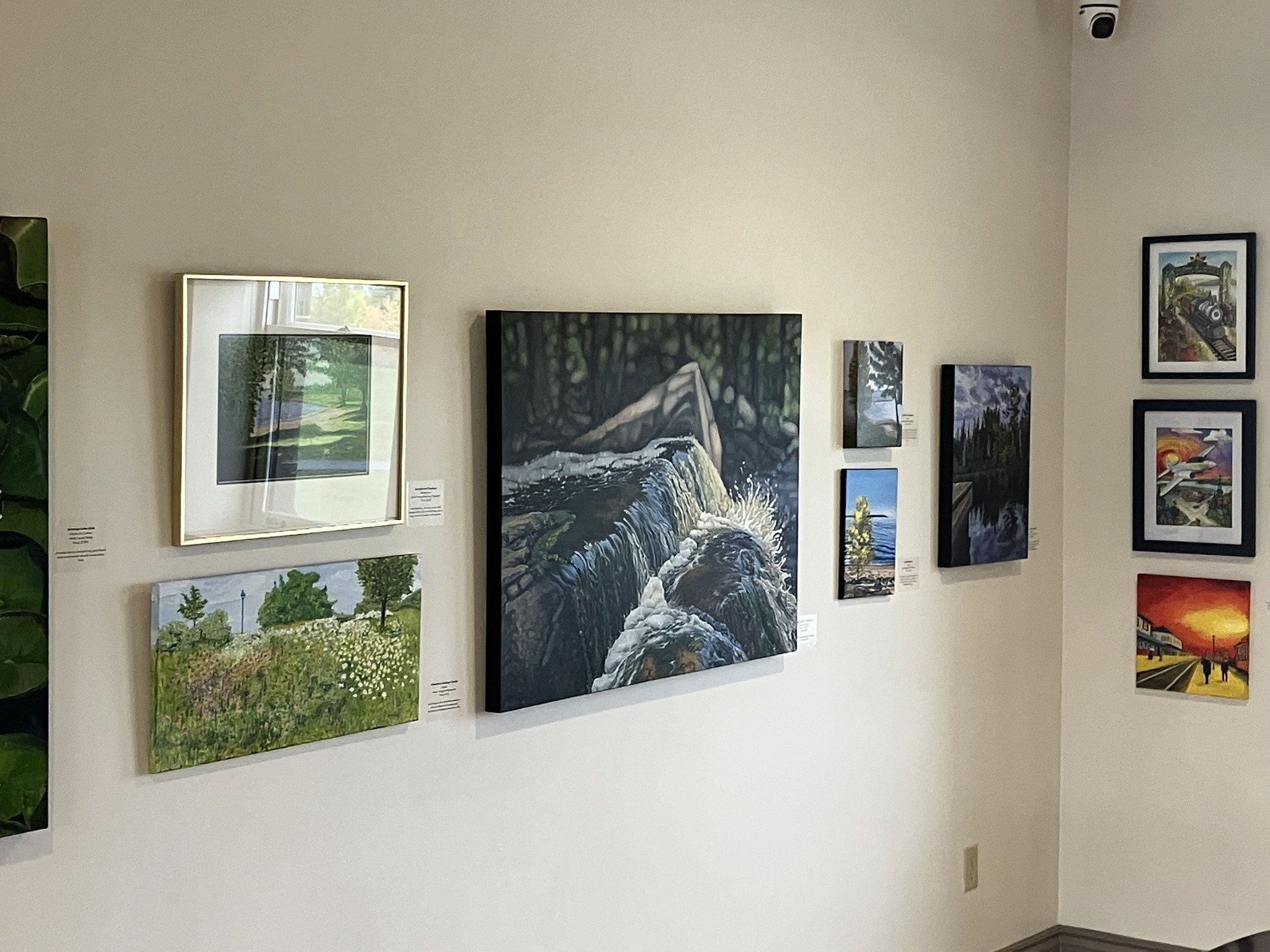 A gallery wall with various landscape paintings including a river scene, lush green fields, trees, and a sunset, displayed in different framed sizes.