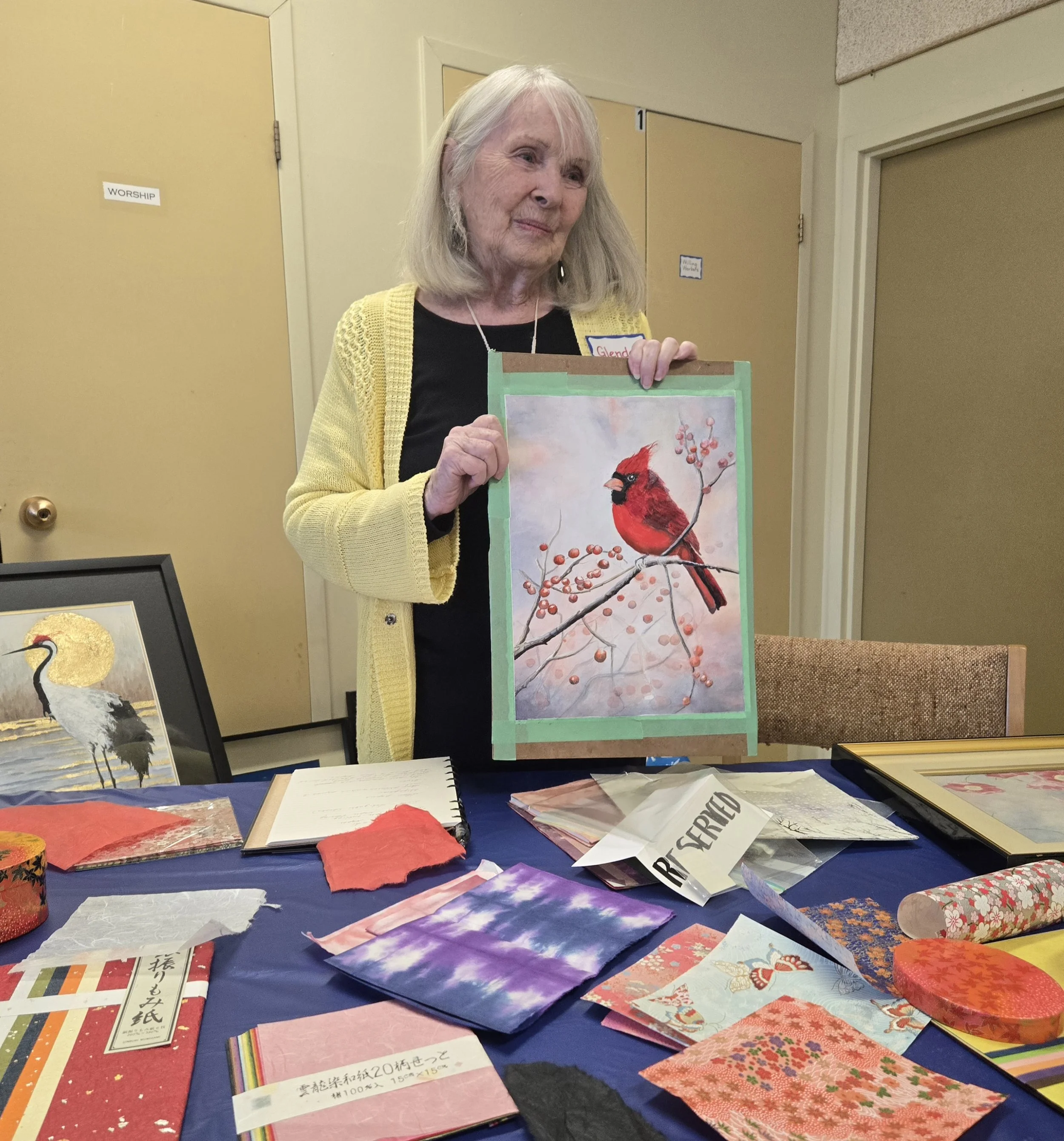 An elderly woman holding a painting of a red cardinal perched on a branch with berries, standing at a table with various fabric and paper crafts, a framed artwork of a crane, and a sign that reads "RESTART" in a room with beige doors and walls.