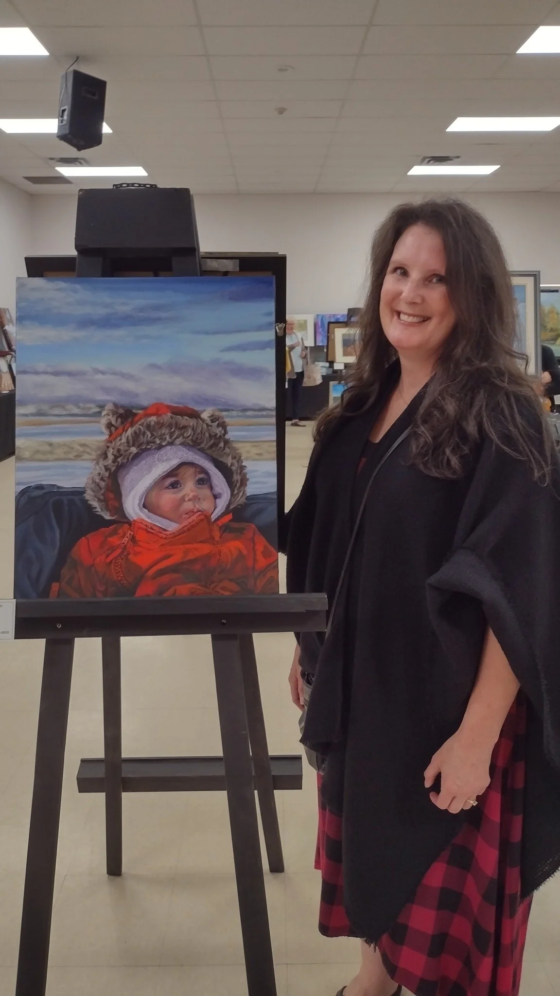 A woman wearing a black jacket and red plaid skirt standing next to a painting of a baby dressed in a red jacket and a furry hood, with a lakeside background and cloudy sky behind them.