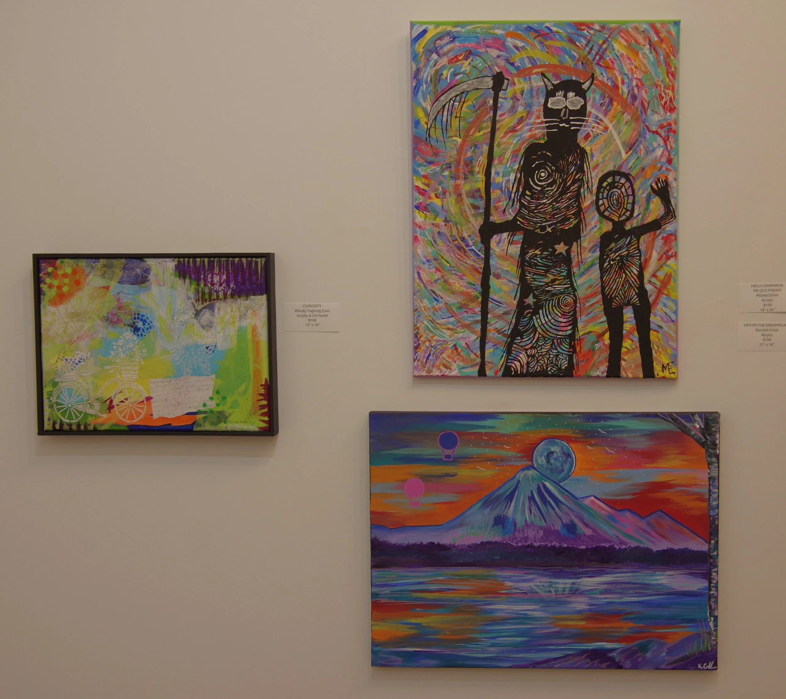 Three colorful paintings are displayed on a white gallery wall. The top right painting features abstract figures of a man with a long staff and a woman, both with spiral designs on their bodies, surrounded by rainbow swirls. The bottom right painting depicts a stylized mountain with a moon and hot air balloons in the sky, above a reflective body of water at sunset. The left painting is more abstract with bright colors and patterns, including some wheel-like shapes.