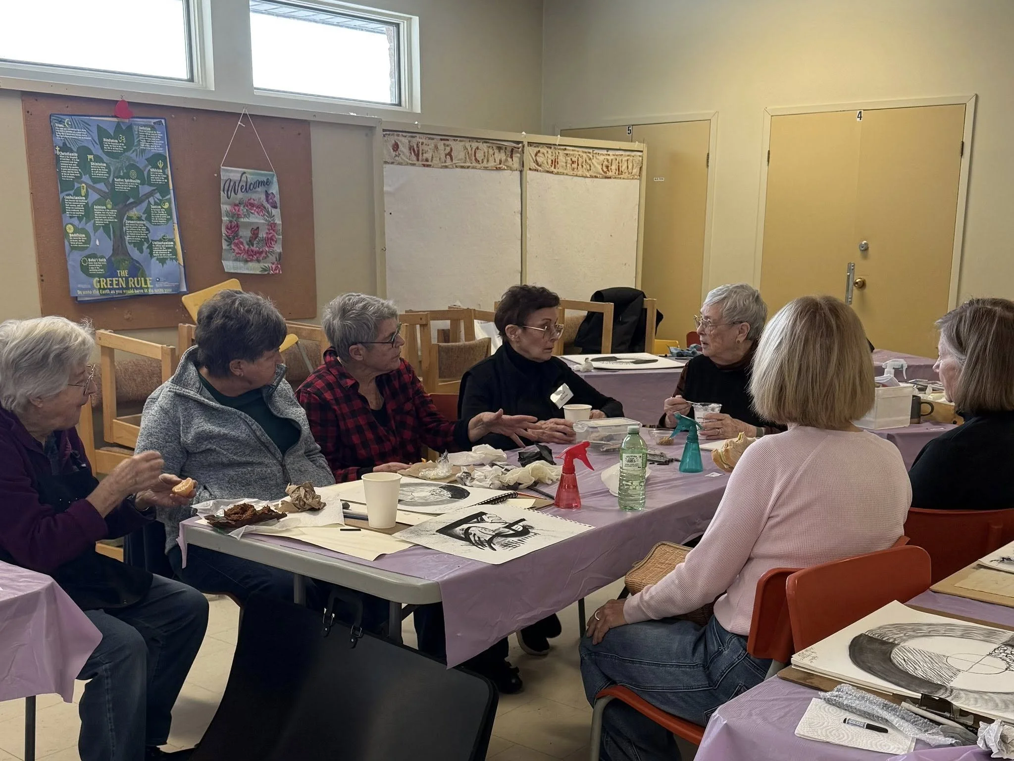 Group of women sitting around a table having a discussion, with food and cleaning spray bottles on the table, in a room with yellow doors and posters on the wall.