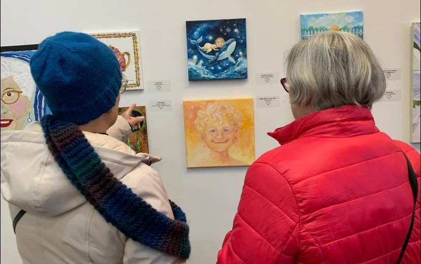 Two women stand in front of a wall with various paintings, observing and discussing one of the artwork pieces. The woman on the left wears a blue knit hat and scarf, and the woman on the right is dressed in a red jacket with gray hair.
